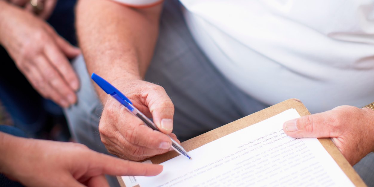 Manos de un cliente que firma un documento en una carpeta de pinza mientras otra mano le señala una cláusula, mejor correduría de seguros Madrid