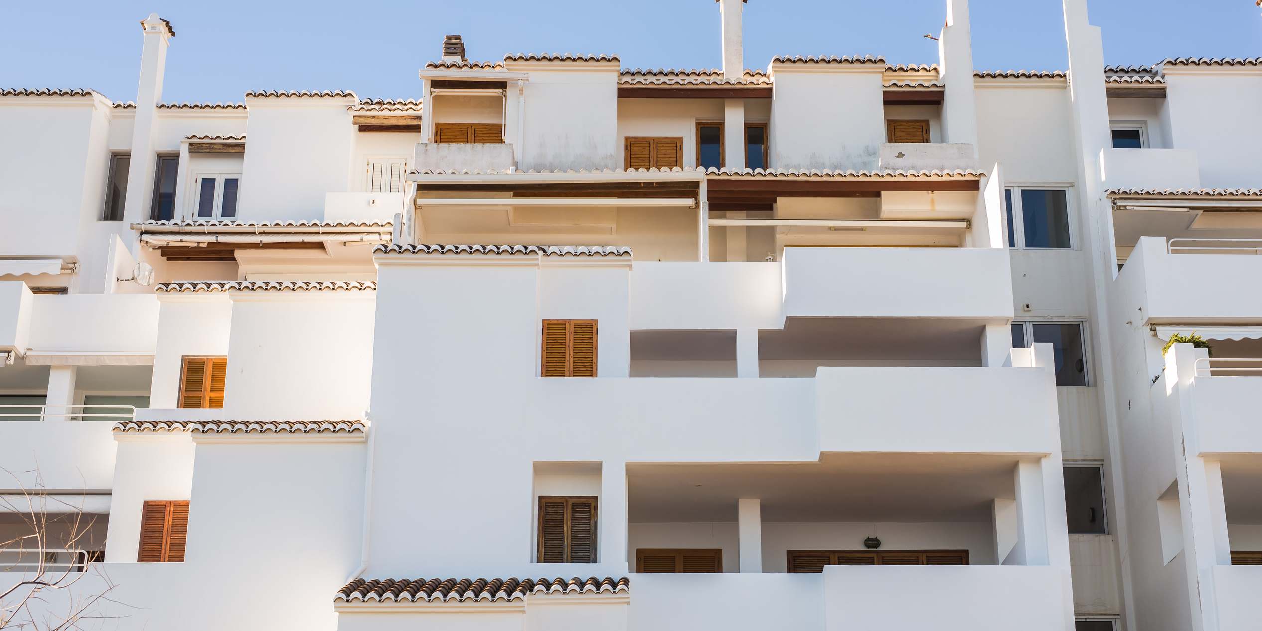 Fachada blanca de un edificio de viviendas con terrazas escalonadas, aleros de teja y contraventanas de madera